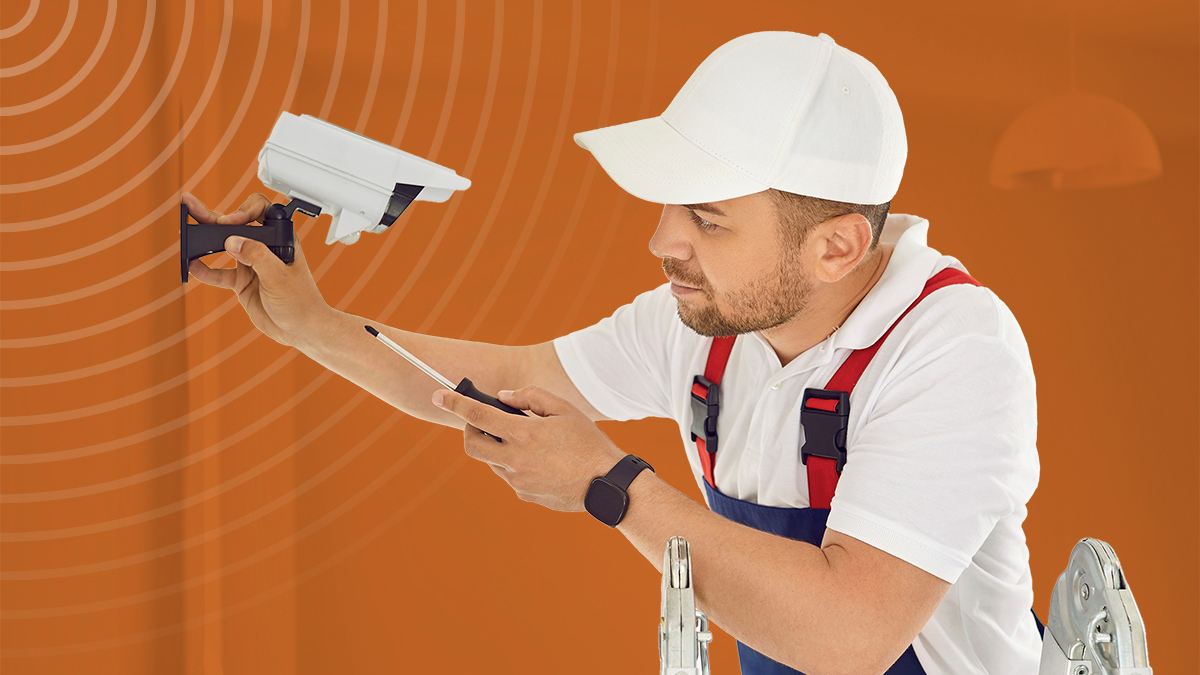 Professional young electrician worker in uniform and cap installs and screws surveillance video camera on wall in bright office space, school, hospital. He is standing on stepladder. Repair works.